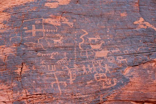 Ancient petroglyphs etched into a reddish-brown rock surface depict various human and animal figures, geometric shapes, and symbols. These carvings suggest storytelling or cultural expressions from a historical civilization.