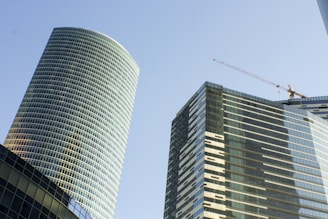 Tall modern skyscrapers with glass facades rise against a clear blue sky. One of the buildings is cylindrical, while the other is rectangular with a crane on its rooftop, indicating construction or maintenance work.