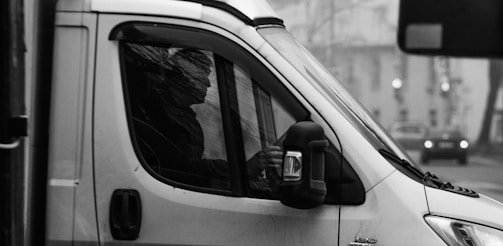 A person is seated inside a vehicle, possibly a van, with their silhouette visible through the window. The scene is in black and white, with a blurred cityscape in the background. The driver's hand is on the steering wheel, suggesting focus and attention on driving.