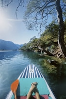 A person is paddleboarding on a serene body of water surrounded by lush greenery and trees. The clear water reflects the bright sky, and a single orange paddle rests on the board. In the distance, mountains can be seen.