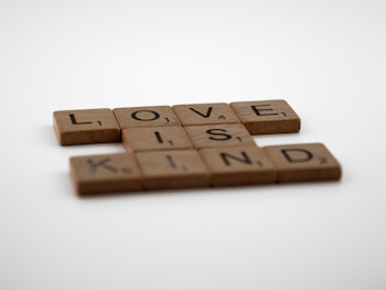 Wooden Scrabble tiles spell out the words 'LOVE IS KIND' against a plain white background. The tiles have a rustic, natural appearance.