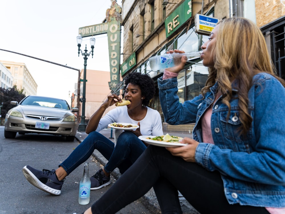 a couple of women sitting on the side of a road eating food