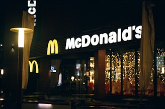 A nighttime view of a McDonald's restaurant with illuminated signage. The entrance is decorated with string lights, creating a warm and inviting ambiance. There are outdoor tables and chairs, and the area is softly lit by lampposts.