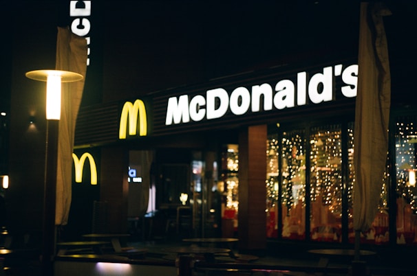 A nighttime view of a McDonald's restaurant with illuminated signage. The entrance is decorated with string lights, creating a warm and inviting ambiance. There are outdoor tables and chairs, and the area is softly lit by lampposts.