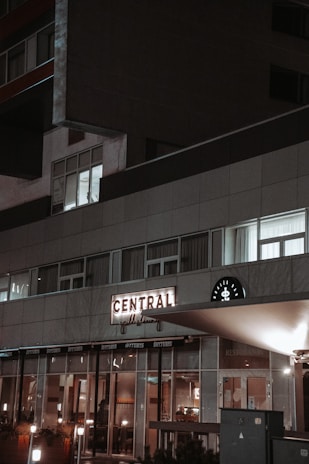 A modern building exterior at night with a prominent illuminated sign reading 'Central' above a grill house entrance. The facade features large windows and a minimalist design. Soft lighting from inside creates a warm, inviting atmosphere.