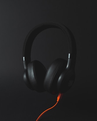 A pair of black over-ear headphones with an orange cable against a dark background. The headphones are centered and appear to be floating, giving a sleek and modern look.