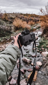 A person holds a digital camera showcasing a scenic view of a rocky stream surrounded by autumn foliage under a cloudy sky. The scene captures a serene outdoor setting with a blend of natural elements.