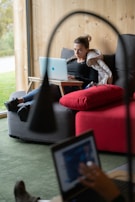 A woman is sitting on a sofa with a wooden wall behind her, working on a laptop placed on a small portable desk. She is relaxed and appears focused on her work. There is a large red cushion on the sofa beside her. Another person, blurred in the foreground, is using a laptop as well.