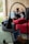 A woman is sitting on a sofa with a wooden wall behind her, working on a laptop placed on a small portable desk. She is relaxed and appears focused on her work. There is a large red cushion on the sofa beside her. Another person, blurred in the foreground, is using a laptop as well.