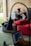 A woman is sitting on a sofa with a wooden wall behind her, working on a laptop placed on a small portable desk. She is relaxed and appears focused on her work. There is a large red cushion on the sofa beside her. Another person, blurred in the foreground, is using a laptop as well.