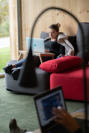 A woman is sitting on a sofa with a wooden wall behind her, working on a laptop placed on a small portable desk. She is relaxed and appears focused on her work. There is a large red cushion on the sofa beside her. Another person, blurred in the foreground, is using a laptop as well.