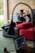 A woman is sitting on a sofa with a wooden wall behind her, working on a laptop placed on a small portable desk. She is relaxed and appears focused on her work. There is a large red cushion on the sofa beside her. Another person, blurred in the foreground, is using a laptop as well.