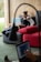 A woman is sitting on a sofa with a wooden wall behind her, working on a laptop placed on a small portable desk. She is relaxed and appears focused on her work. There is a large red cushion on the sofa beside her. Another person, blurred in the foreground, is using a laptop as well.