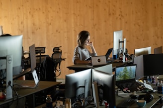 A woman is seated in a modern office setting, surrounded by multiple computer monitors and laptops on a desk. The room has a wooden wall paneling that lends a warm ambiance. The workspace includes chairs and various office supplies, indicating an active work environment.
