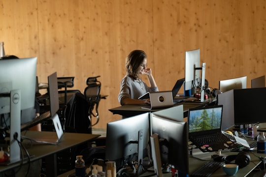 A woman is seated in a modern office setting, surrounded by multiple computer monitors and laptops on a desk. The room has a wooden wall paneling that lends a warm ambiance. The workspace includes chairs and various office supplies, indicating an active work environment.