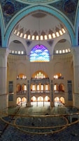 A grand interior of a mosque featuring an intricate dome adorned with ornate patterns and calligraphy. Stained glass windows filter colorful light into the room, illuminating its vast expanse. The floor is covered with a richly patterned carpet, and a large chandelier hangs prominently in the center. The architectural details display Islamic design elements.