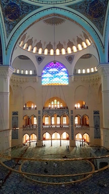 A grand interior of a mosque featuring an intricate dome adorned with ornate patterns and calligraphy. Stained glass windows filter colorful light into the room, illuminating its vast expanse. The floor is covered with a richly patterned carpet, and a large chandelier hangs prominently in the center. The architectural details display Islamic design elements.