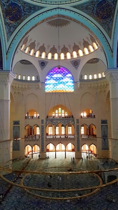 A grand interior of a mosque featuring an intricate dome adorned with ornate patterns and calligraphy. Stained glass windows filter colorful light into the room, illuminating its vast expanse. The floor is covered with a richly patterned carpet, and a large chandelier hangs prominently in the center. The architectural details display Islamic design elements.
