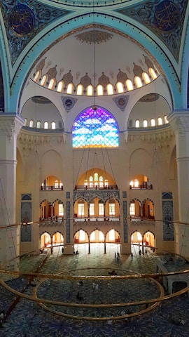 A grand interior of a mosque featuring an intricate dome adorned with ornate patterns and calligraphy. Stained glass windows filter colorful light into the room, illuminating its vast expanse. The floor is covered with a richly patterned carpet, and a large chandelier hangs prominently in the center. The architectural details display Islamic design elements.