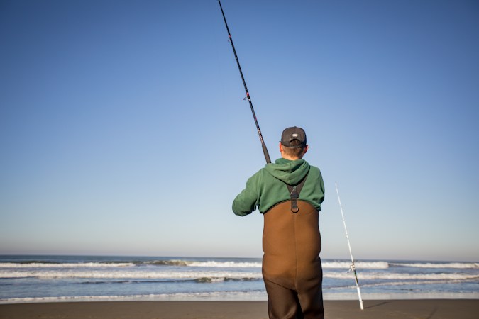 Surf angler seen from behind in brown chest waders with suspenders and a green hoodie, holding a long surf rod up, a second rod in a sand spike beside him and low waves ahead
