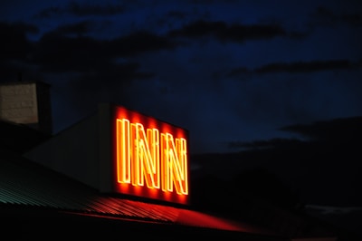 A brightly illuminated neon sign with the word 'INN' stands out against a dark, nighttime sky. The sign emits a warm orange-red glow, contrasting with the silhouetted outlines of nearby buildings and clouds.