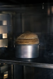 A loaf of bread rising in a metallic pan inside an oven with a dimly lit interior.
