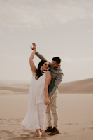 A couple enjoying a romantic moment in a desert setting with sand dunes in the background. The woman is wearing a flowing white dress, and the man is dressed in a grey shirt and beige pants. They are barefoot, and the man appears to be guiding the woman's dance with a tender gesture.