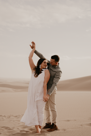 A couple enjoying a romantic moment in a desert setting with sand dunes in the background. The woman is wearing a flowing white dress, and the man is dressed in a grey shirt and beige pants. They are barefoot, and the man appears to be guiding the woman's dance with a tender gesture.