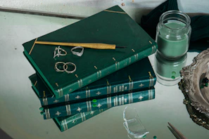 A stack of books with golden spines next to a green forest-themed notebook and a silver pen