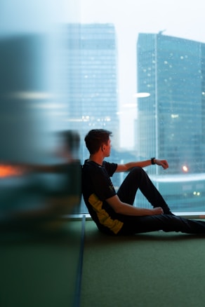 A young person is sitting indoors by a large window, gazing outside. The view through the window shows modern urban architecture with tall buildings. The indoor environment is dimly lit, creating a contrast with the bright city lights visible through the glass.