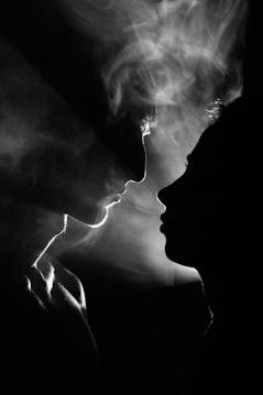 Two human profiles are shown in silhouette against a smoky background, with the faces almost touching. The use of black and white creates a dramatic play of light and shadow, emphasizing the outlines and creating a mysterious atmosphere.