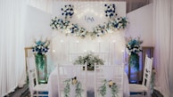 An elegantly decorated indoor space features a dining setup with white chairs adorned with green foliage and white flowers. A backdrop with white and blue floral arrangements and subtle lighting creates a romantic atmosphere. The initials 'L & A' are displayed on the backdrop, indicating a personalized event. Tall floral arrangements flank the setup, complementing the theme.