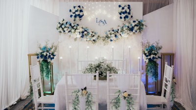 An elegantly decorated indoor space features a dining setup with white chairs adorned with green foliage and white flowers. A backdrop with white and blue floral arrangements and subtle lighting creates a romantic atmosphere. The initials 'L & A' are displayed on the backdrop, indicating a personalized event. Tall floral arrangements flank the setup, complementing the theme.