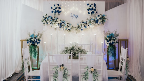 An elegantly decorated indoor space features a dining setup with white chairs adorned with green foliage and white flowers. A backdrop with white and blue floral arrangements and subtle lighting creates a romantic atmosphere. The initials 'L & A' are displayed on the backdrop, indicating a personalized event. Tall floral arrangements flank the setup, complementing the theme.