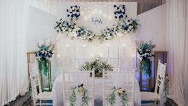 An elegantly decorated indoor space features a dining setup with white chairs adorned with green foliage and white flowers. A backdrop with white and blue floral arrangements and subtle lighting creates a romantic atmosphere. The initials 'L & A' are displayed on the backdrop, indicating a personalized event. Tall floral arrangements flank the setup, complementing the theme.