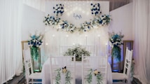 An elegantly decorated indoor space features a dining setup with white chairs adorned with green foliage and white flowers. A backdrop with white and blue floral arrangements and subtle lighting creates a romantic atmosphere. The initials 'L & A' are displayed on the backdrop, indicating a personalized event. Tall floral arrangements flank the setup, complementing the theme.