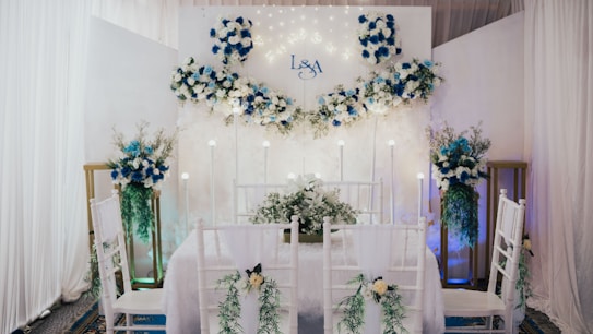 An elegantly decorated indoor space features a dining setup with white chairs adorned with green foliage and white flowers. A backdrop with white and blue floral arrangements and subtle lighting creates a romantic atmosphere. The initials 'L & A' are displayed on the backdrop, indicating a personalized event. Tall floral arrangements flank the setup, complementing the theme.