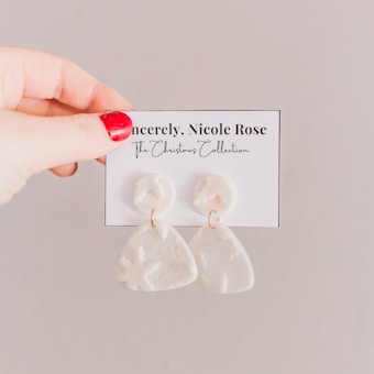 A hand with red-painted nails holding a card displaying a pair of white, triangular dangle earrings. The card has text 'Sincerely, Nicole Rose' and 'The Christmas Collection' written on it.