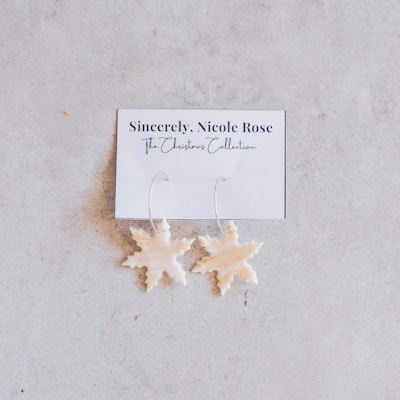 A pair of white, snowflake-shaped earrings attached to a card labeled 'Sincerely, Nicole Rose - The Christmas Collection.' They are placed against a light, neutral-colored background.