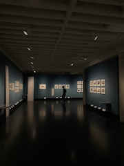 A dimly lit art gallery with dark blue walls, featuring framed artworks arranged neatly. A lone figure is seen walking through the gallery, casting a reflection on the polished floor. Spotlights from the ceiling illuminate the artwork, creating a focused ambiance.