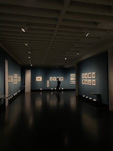 A dimly lit art gallery with dark blue walls, featuring framed artworks arranged neatly. A lone figure is seen walking through the gallery, casting a reflection on the polished floor. Spotlights from the ceiling illuminate the artwork, creating a focused ambiance.