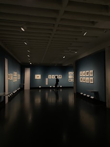 A dimly lit art gallery with dark blue walls, featuring framed artworks arranged neatly. A lone figure is seen walking through the gallery, casting a reflection on the polished floor. Spotlights from the ceiling illuminate the artwork, creating a focused ambiance.