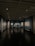 A dimly lit art gallery with dark blue walls, featuring framed artworks arranged neatly. A lone figure is seen walking through the gallery, casting a reflection on the polished floor. Spotlights from the ceiling illuminate the artwork, creating a focused ambiance.