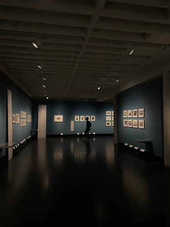 A dimly lit art gallery with dark blue walls, featuring framed artworks arranged neatly. A lone figure is seen walking through the gallery, casting a reflection on the polished floor. Spotlights from the ceiling illuminate the artwork, creating a focused ambiance.
