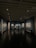 A dimly lit art gallery with dark blue walls, featuring framed artworks arranged neatly. A lone figure is seen walking through the gallery, casting a reflection on the polished floor. Spotlights from the ceiling illuminate the artwork, creating a focused ambiance.