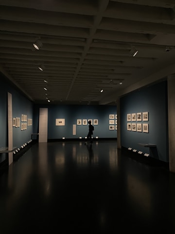 A dimly lit art gallery with dark blue walls, featuring framed artworks arranged neatly. A lone figure is seen walking through the gallery, casting a reflection on the polished floor. Spotlights from the ceiling illuminate the artwork, creating a focused ambiance.