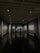 A dimly lit art gallery with dark blue walls, featuring framed artworks arranged neatly. A lone figure is seen walking through the gallery, casting a reflection on the polished floor. Spotlights from the ceiling illuminate the artwork, creating a focused ambiance.