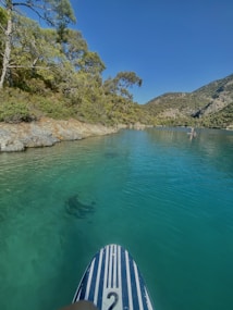 A peaceful body of turquoise water surrounded by rocky and forested hills. A paddle board with a striped design is in the foreground, and a person is paddleboarding further in the distance. Overhanging trees and shrubs add to the natural scenery.