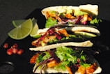 Three tacos filled with grilled chicken, sliced avocado, fresh cilantro, and red cabbage are arranged on a dark surface. The tacos are garnished with lime wedges and small red berries.
