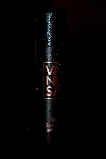 A sleek black cylindrical spray can with the word 'VANESA' printed vertically in copper lettering. The can features a subtle butterfly logo and the words 'SHER' and 'BODY DE'. Water droplets are visible on the surface, adding a dynamic, fresh feel.
